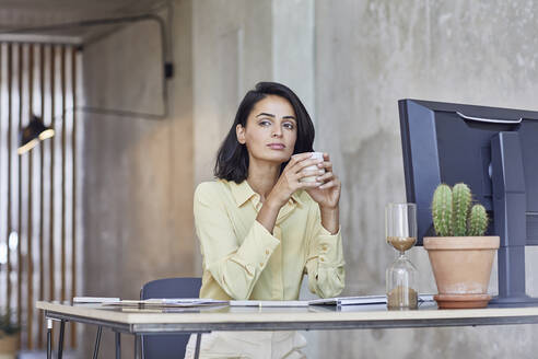 Nachdenkliche Geschäftsfrau, die eine Kaffeetasse hält, während sie am Schreibtisch im Büro sitzt - MCF01232