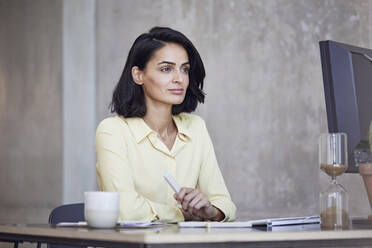 Thoughtful female entrepreneur looking away while sitting at desk against wall in office - MCF01234