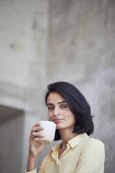 Nahaufnahme einer selbstbewussten Geschäftsfrau, die eine Kaffeetasse an die Wand eines Büros hält - MCF01244
