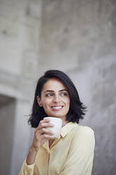 Close-up of smiling female entrepreneur holding coffee cup against wall in office - MCF01245