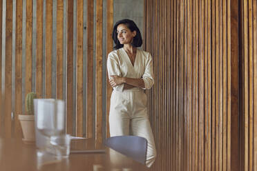 Smiling female entrepreneur with arms crossed standing against wooden doors in office - MCF01253