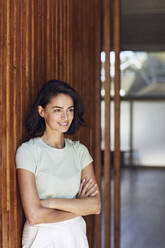 Thoughtful female entrepreneur with arms crossed standing by wooden wall in office - MCF01261