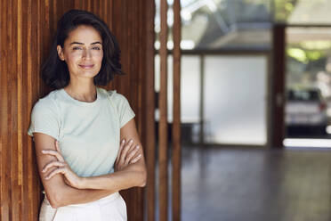 Smiling female entrepreneur with arms crossed standing by wooden wall in office - MCF01262