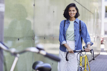 Smiling businesswoman with bicycle standing by wall in city - MCF01267