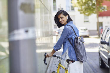 Smiling female commuter with backpack holding bicycle while standing in city - MCF01274