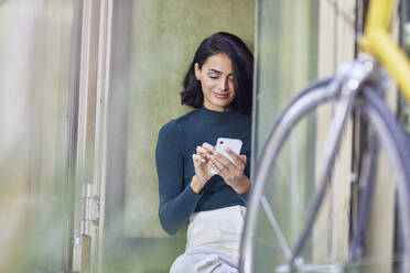 Lächelnde Geschäftsfrau, die ein Smartphone benutzt, während sie an der Tür im Büro sitzt - MCF01280