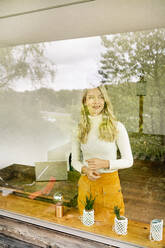 Smiling businesswoman looking away while standing by window in office seen through glass - PESF02067