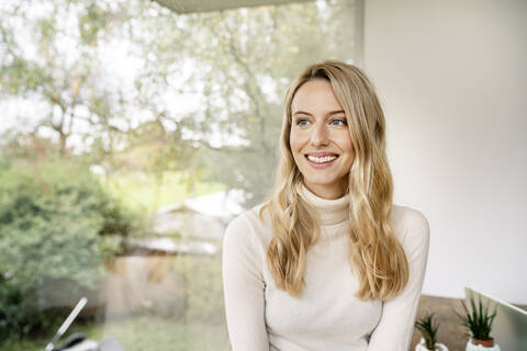 Smiling thoughtful businesswoman with blond hair sitting by window in office stock photo