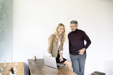 Lächelnde Geschäftsfrau, die mit einem männlichen Kollegen an der Wand im Büro über einen Laptop diskutiert - PESF02102