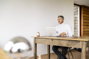 Nachdenklicher Geschäftsmann mit Laptop auf dem Schreibtisch sitzend an der Wand im Büro - PESF02111
