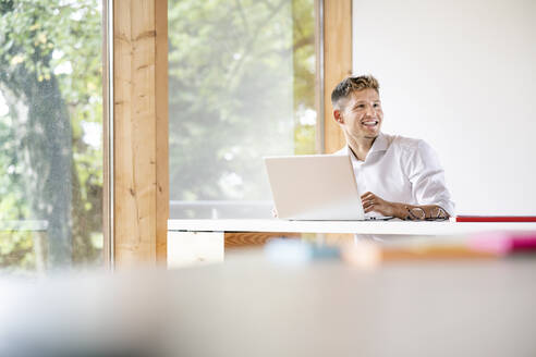 Lächelnder Geschäftsmann mit Laptop auf dem Schreibtisch, der wegschaut, während er am Fenster im Büro sitzt - PESF02124