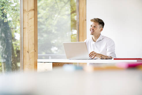 Lächelnder Geschäftsmann mit Laptop auf dem Schreibtisch, der im Büro sitzt und nachdenkt - PESF02125