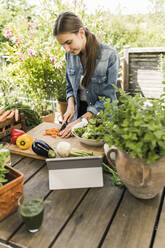 Young woman with digital tablet on table chopping vegetables in yard - UUF21002