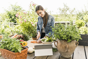 Junge Frau, die beim Gemüseschneiden im Garten auf ein digitales Tablet mit Rezept schaut - UUF21003