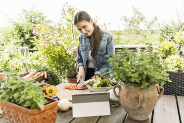 Young woman looking recipe over digital tablet while chopping vegetables in yard - UUF21003