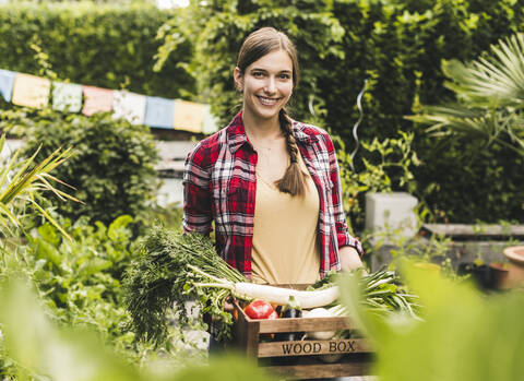 Lächelnde Frau, die Gemüse in einer Kiste trägt, während sie an Pflanzen im Garten steht, lizenzfreies Stockfoto