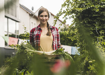 Lächelnde Frau, die Bio-Gemüse in einer Kiste trägt, während sie im Garten steht - UUF21010