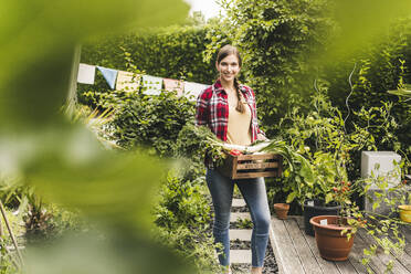 Smiling beautiful woman with vegetables in crate standing against plants at garden - UUF21011