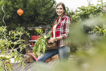 Lächelnde junge Frau, die eine Kiste trägt, während sie im Gemüsegarten steht - UUF21012