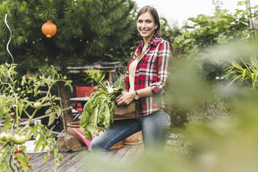 Smiling young woman carrying crate while standing in vegetable garden - UUF21012