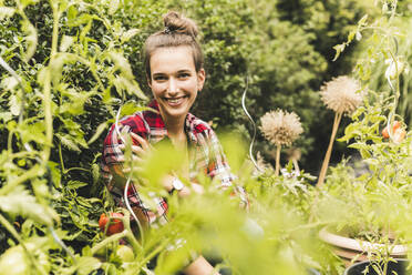 Smiling beautiful woman sitting amidst plants in vegetable garden - UUF21017