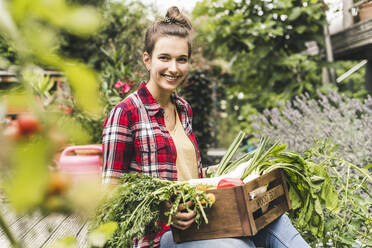 Smiling beautiful woman with vegetable crate sitting in community garden - UUF21019