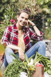 Lächelnde schöne Frau mit Gemüse im Gemeinschaftsgarten sitzend - UUF21021