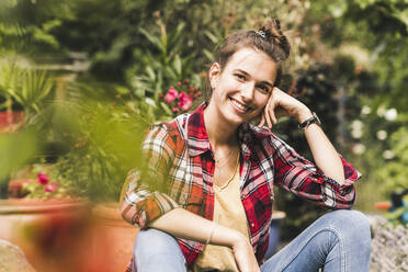 Smiling beautiful woman sitting against plants in vegetable garden - UUF21022