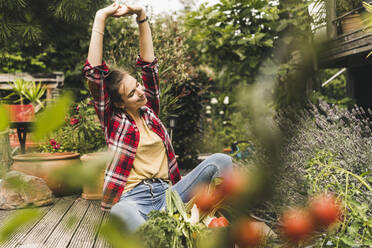 Young woman with arms raised sitting against plants in vegetable garden - UUF21023