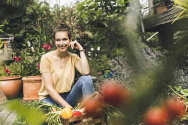 Beautiful woman with vegetables sitting amidst plants in community garden - UUF21024