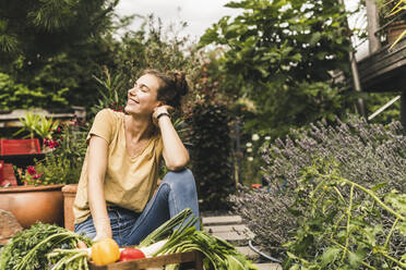 Young woman with eyes closed sitting by vegetables and plants in community garden - UUF21025