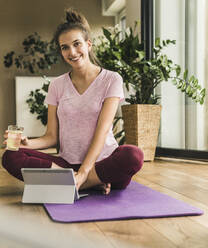 Smiling young woman with water and digital tablet sitting on exercise mat at home - UUF21029