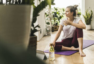 Thoughtful young woman holding drink while sitting on mat at home - UUF21034