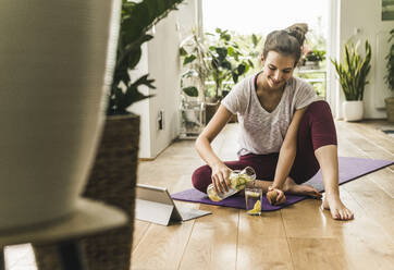 Smiling young woman pouring drink in glass while sitting on exercise mat at home - UUF21035