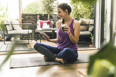 Smiling young woman holding drink while using digital tablet on exercise mat at home - UUF21039