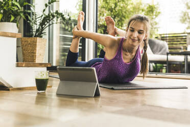 Junge Frau, die in ein digitales Tablet schaut, während sie zu Hause auf einer Matte Yoga übt - UUF21044