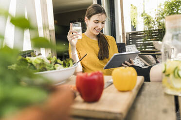 Lächelnde junge Frau mit Gemüse auf dem Tisch, die zu Hause ein Video über ein digitales Tablet ansieht - UUF21048