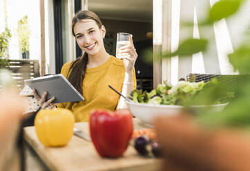 Lächelnde junge Frau hält ein digitales Tablet und trinkt ein Glas, während sie zu Hause am Tisch sitzt - UUF21049