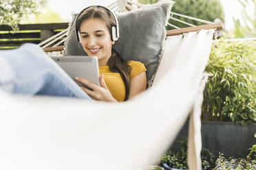 Lächelnde junge Frau mit Kopfhörern, die ein digitales Tablet benutzt, während sie sich in einer Hängematte im Garten entspannt - UUF21054