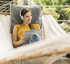 Lächelnde Frau mit digitalem Tablet, die wegschaut, während sie auf einer Hängematte im Garten liegt - UUF21055