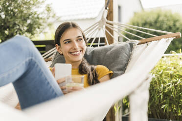 Thoughtful smiling woman with smart phone relaxing on hammock - UUF21061