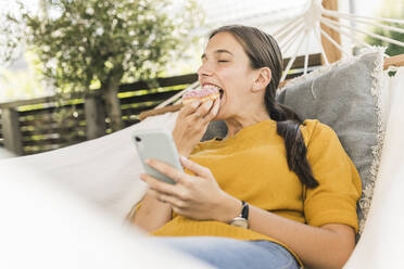 Junge Frau isst Donut, während sie ihr Smartphone in der Hängematte im Garten benutzt - UUF21071