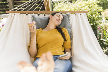 Junge Frau mit geschlossenen Augen, die einen Donut und ein Smartphone hält, während sie sich in der Hängematte entspannt - UUF21072