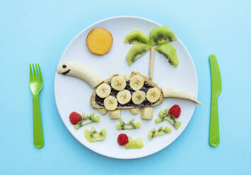 Frühstück mit Dinosaurierform aus Banane, Kiwi, Brot, Schokoladenaufstrich und Beeren auf weißem Teller auf blauem Hintergrund - GEMF04126