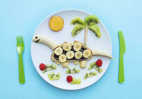 Breakfast with dinosaur shape made of banana, kiwi, bread, chocolate spread and berries on white plate on blue background - GEMF04126