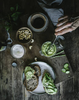 Aus der Hand einer nicht erkennbaren Person, die auf einem Holzbrett Toasts mit grüner Cashewpastete und Gurkenscheiben zubereitet - ADSF14312