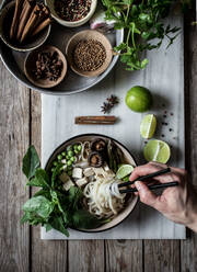 From above person hands with arranged bowls with dry spices and served Pho soup with noodles on marble board - ADSF14334