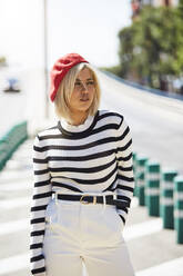 Junge blonde Frau in gestreiftem schwarz-weißem Hemd und roter französischer Mütze geht auf unscharfem Stadthintergrund - ADSF14342