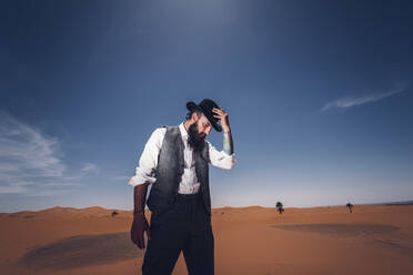 Bärtiger Mann im Cowboykostüm, der in der Wüste vor blauem Himmel steht und nach unten schaut - ADSF14347