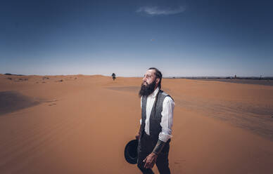 Bärtiger Mann im Cowboy-Kostüm schaut weg, während er in der Wüste vor blauem Himmel steht - ADSF14348